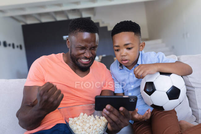 Afrikanisch-amerikanischer Vater und Sohn sitzen auf dem Sofa und bedienen sich eines Smartphones. Zuhause in Isolation während der Quarantäne. — Stockfoto