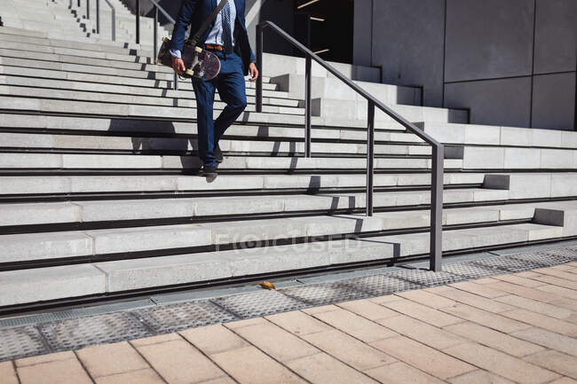 Low section of businessman holding skateboard, walking downstairs in the sun. hanging out at an urban skatepark in summer. — Stock Photo