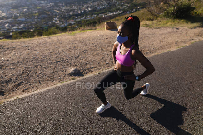 Fit african american woman in face mask stretching exercising in countryside. healthy active lifestyle and outdoor fitness during coronavirus covid 19 pandemic. — Stock Photo