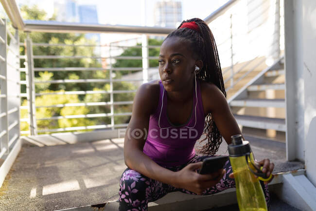Ajuste mujer afroamericana sentada en escalones con auriculares usando teléfono inteligente durante el ejercicio en la ciudad. estilo de vida activo urbano saludable y fitness al aire libre. - foto de stock