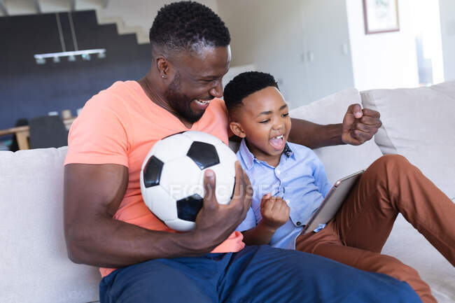 African american father and son sitting on sofa, using tablet and smiling. at home in isolation during quarantine lockdown. — Stock Photo