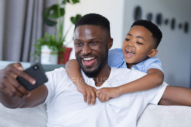 African american father and son sitting on sofa, using smartphone and smiling. at home in isolation during quarantine lockdown. — Stock Photo