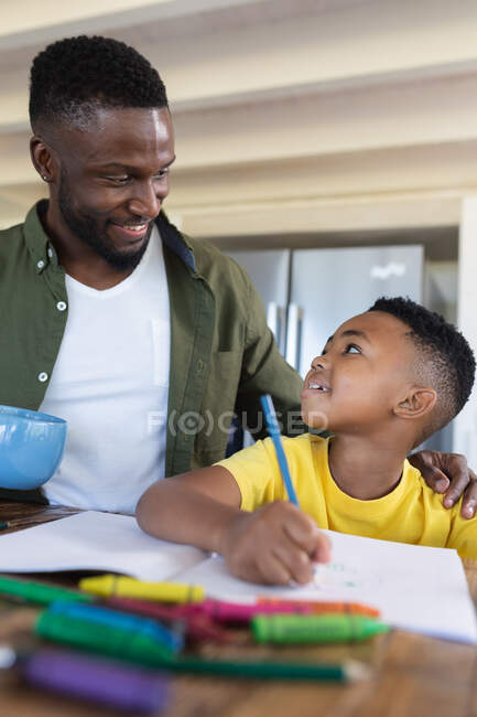 Padre e hijo afroamericanos sentados en la mesa, escribiendo en cuaderno sonriendo en casa aislados durante el bloqueo de cuarentena. - foto de stock
