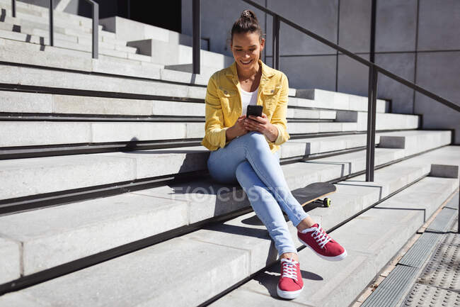 Усміхнена біла жінка сидить на сходах і використовує смартфон. висить у міському скейтпарку влітку . — стокове фото