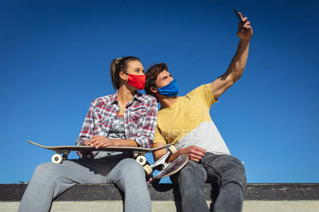 Белая женщина и мужчина в масках, сидят на стене и делают селфи. тусоваться в городском скейтпарке летом во время пандемии коронавируса ковид 19. — стоковое фото