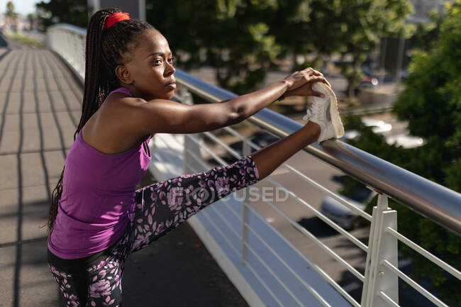 Fit african american woman stretching leg on foot bridge exercising in city. healthy active lifestyle and outdoor fitness. — Stock Photo