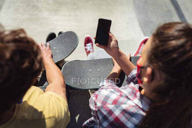 Caucasian male and female friends wearing face masks sitting with skateboards, using smartphone. hanging out at urban skatepark in summer during coronavirus covid 19 pandemic. — Stock Photo