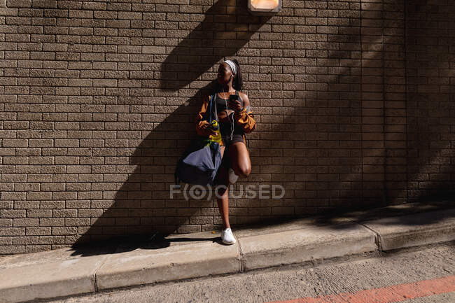 Fit african american woman with gym bag and water bottle leaning against brick wall in city. healthy urban active lifestyle and outdoor fitness. — Stock Photo