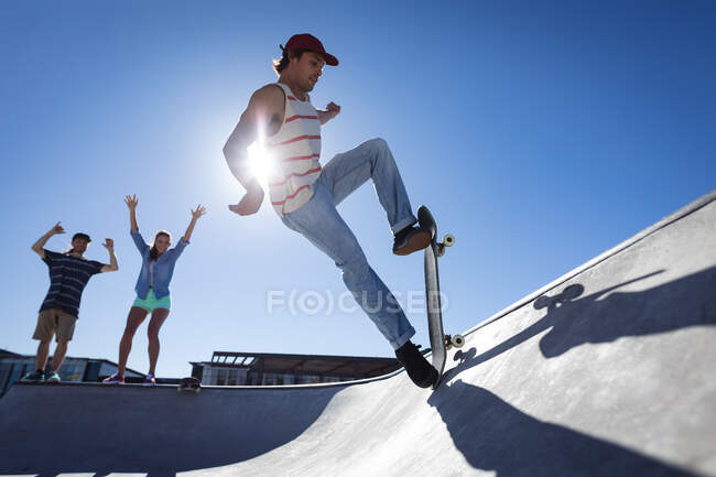 Caucasian man skateboarding on sunny day while two friends cheer leading him. hanging out at urban skatepark in summer. — Stock Photo