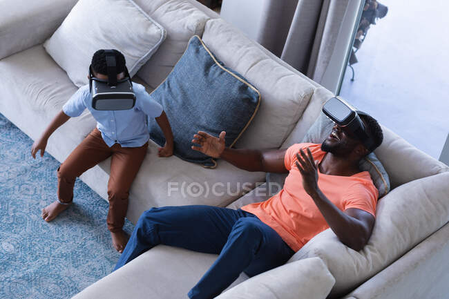 Padre e hijo afroamericanos sentados en el sofá, con auriculares vr tocando la pantalla virtual en casa en aislamiento durante el bloqueo de cuarentena. - foto de stock