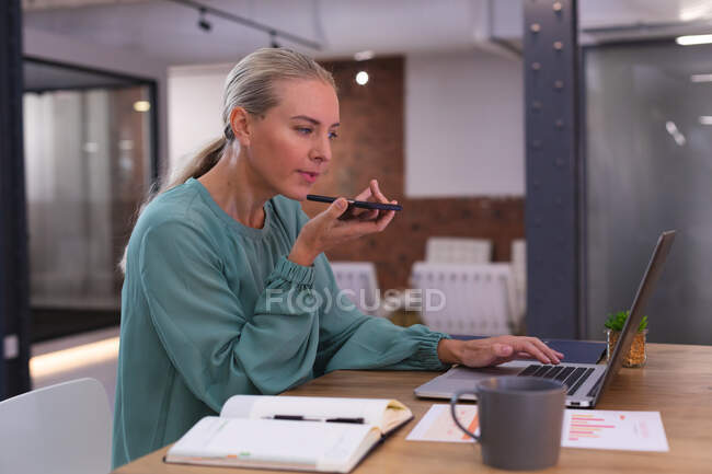 Mulher caucasiana falando no smartphone enquanto usa laptop no escritório moderno. negócio, profissionalismo e conceito de escritório — Fotografia de Stock