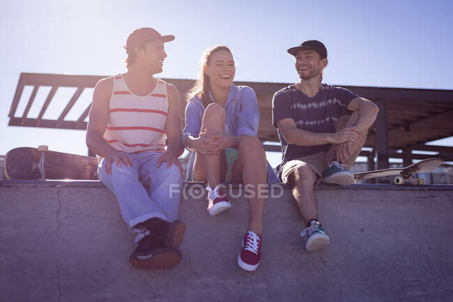Three happy caucasian female and male friends sitting on wall and laughing in the sun. hanging out at an urban skatepark in summer. — Stock Photo