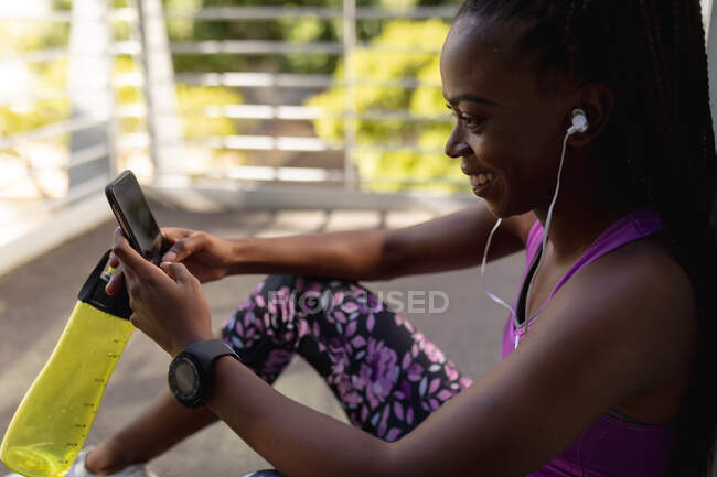 Sonriendo ajuste mujer afroamericana sentada con auriculares usando smartphone durante el ejercicio en la ciudad. estilo de vida activo urbano saludable y fitness al aire libre. - foto de stock