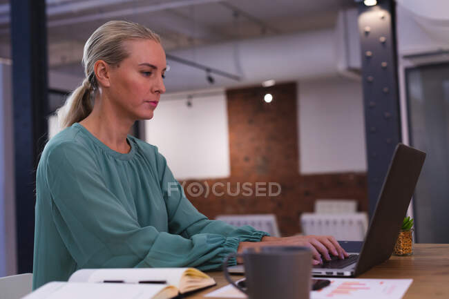 Mujer joven caucásica que usa el ordenador portátil mientras trabaja en la oficina moderna. negocio, profesionalidad y concepto de oficina - foto de stock