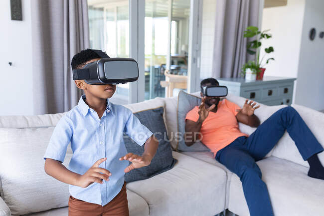 African american father and son sitting on sofa, wearing vr headset touching virtual screen at home in isolation during quarantine lockdown. — Stock Photo