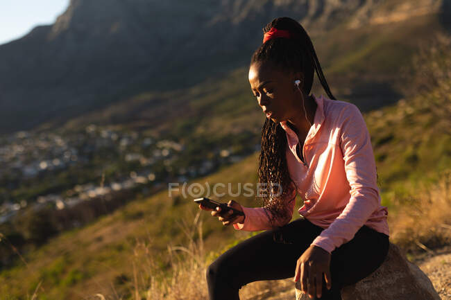 African american woman taking break in exercise outdoors, wearing earphones and using smartphone. healthy active lifestyle and outdoor fitness. — Stock Photo