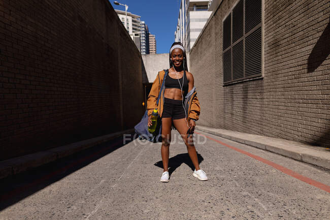 Sorrindo apto mulher afro-americana com saco de ginástica e garrafa de água em pé na rua na cidade. estilo de vida ativo urbano saudável e aptidão ao ar livre. — Fotografia de Stock