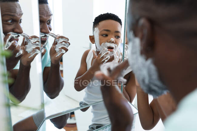 Afro-americano pai e filho no banheiro, aplicando espuma de barbear. em casa em isolamento durante o confinamento de quarentena. — Fotografia de Stock