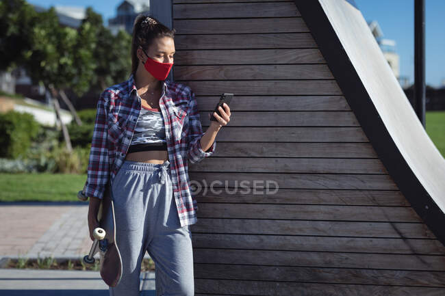 Donna caucasica indossando maschera facciale, tenendo lo skateboard e utilizzando smartphone. appendere fuori a skatepark urbano in estate durante coronavirus covid 19 pandemia. — Foto stock