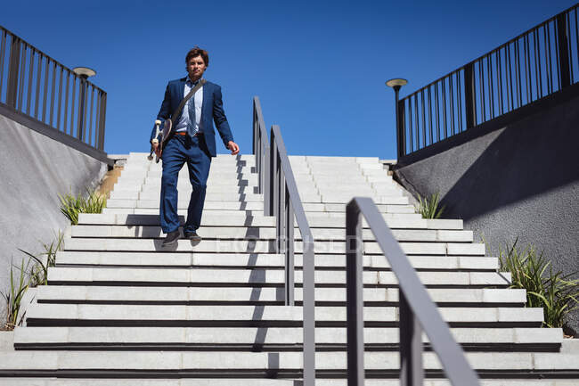 Caucasian businessman holding skateboard, walking downstairs in the sun. hanging out at an urban skatepark in summer. — Stock Photo