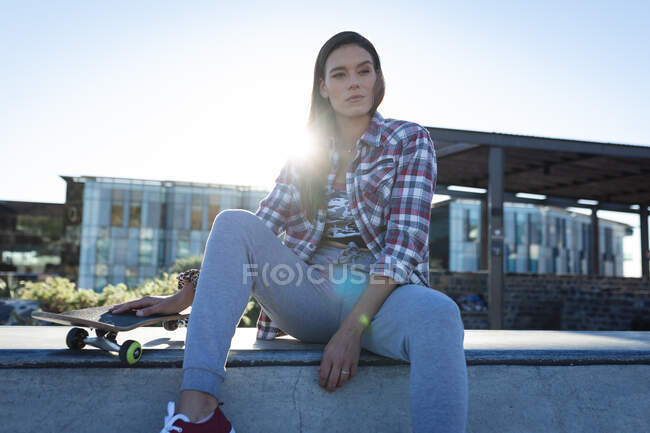 Retrato de mulher caucasiana sentada na parede com skate ao sol. sair em um parque de skate urbano no verão. — Fotografia de Stock