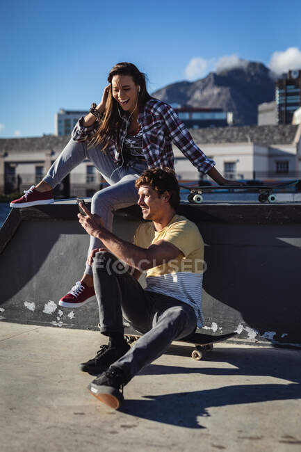 Mulher caucasiana feliz e homem sentado na parede com skates, usando smartphone ao sol. sair em um parque de skate urbano no verão. — Fotografia de Stock
