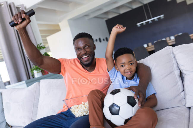 Padre e hijo afroamericanos sentados en un sofá, viendo televisión y sonriendo en casa aislados durante el cierre de cuarentena. - foto de stock