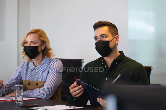 Colegas caucásicas de sexo masculino y femenino con máscaras faciales sentadas en la sala de reuniones de la oficina moderna. higiene y distanciamiento social en el lugar de trabajo durante la pandemia covid 19. - foto de stock