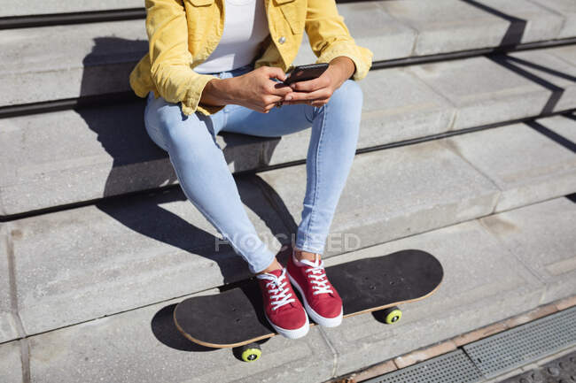Sección baja de mujer caucásica sentada en escaleras con monopatín y usando smartphone. pasando el rato en skatepark urbano en verano. - foto de stock