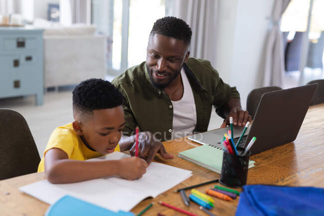Père et fils afro-américains assis à table, en utilisant un ordinateur portable et en écrivant dans un carnet. à domicile en isolement pendant le confinement en quarantaine. — Photo de stock