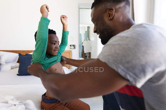 African american father and son sitting on bed, helping with dressing up. at home in isolation during quarantine lockdown. — Stock Photo