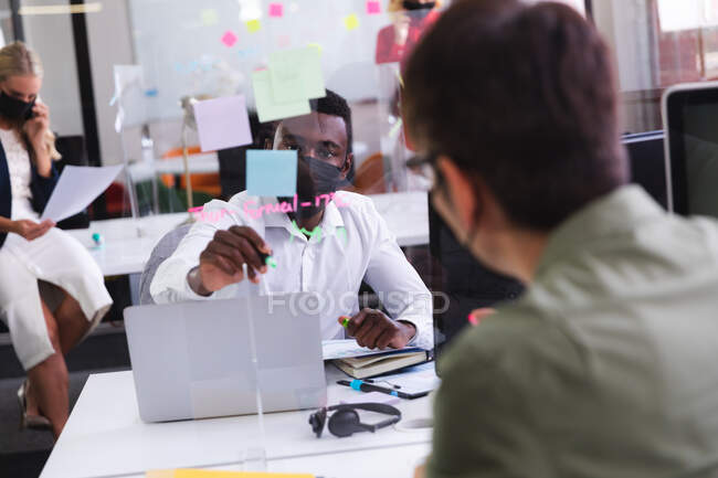 Uomo afroamericano indossando maschera viso scrittura su tavola di vetro mentre seduto sulla scrivania in ufficio. igiene e distanza sociale sul posto di lavoro durante la covd 19 pandemia. — Foto stock