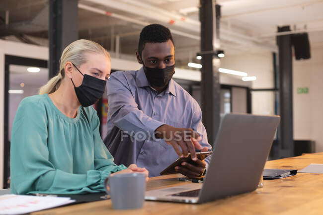 Різні чоловічі та жіночі офісні колеги в масках для обличчя обговорюють ноутбук в сучасному офісі. гігієна та соціальне дистанціювання на робочому місці під час пандемії ковадла-19 . — стокове фото