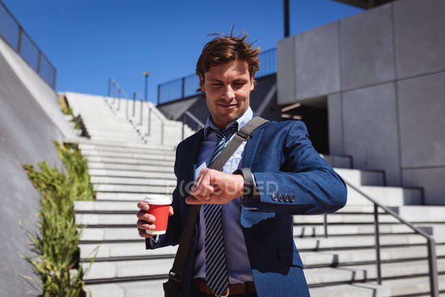 Un uomo d'affari caucasico che tiene il caffe 'e guarda lo smartwatch al sole. uscire in estate in uno skatepark urbano. — Foto stock