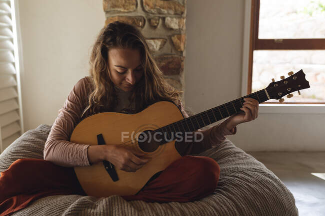 Felice donna caucasica seduta sul sacchetto di fagioli a suonare la chitarra acustica nel soleggiato soggiorno cottage. semplice vivere in una casa rurale fuori dalla griglia. — Foto stock