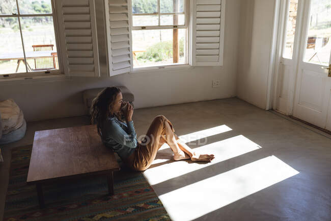 Kaukasische Frau sitzt auf dem Boden und trinkt Kaffee im sonnigen Wohnzimmer der Hütte. einfaches Leben in einer netzunabhängigen ländlichen Wohnung. — Stockfoto
