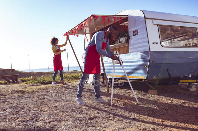Diversa pareja abriendo y preparando camión de comida junto al mar en el día soleado. concepto de empresa independiente y servicio de comida callejera. - foto de stock