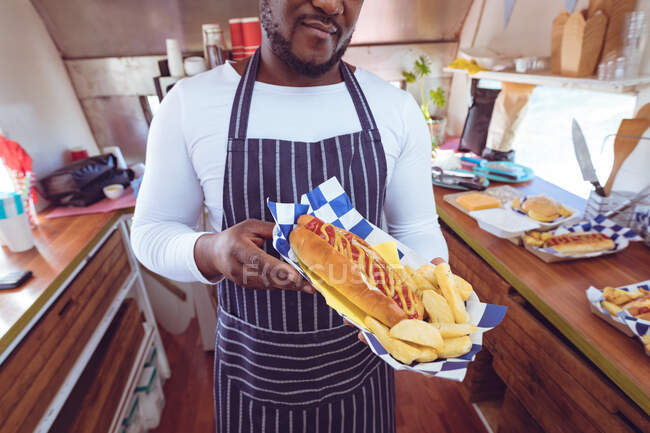 Midsection of african american man in food truck holding hot-dog and chips. independent business and street food service concept. — Stock Photo