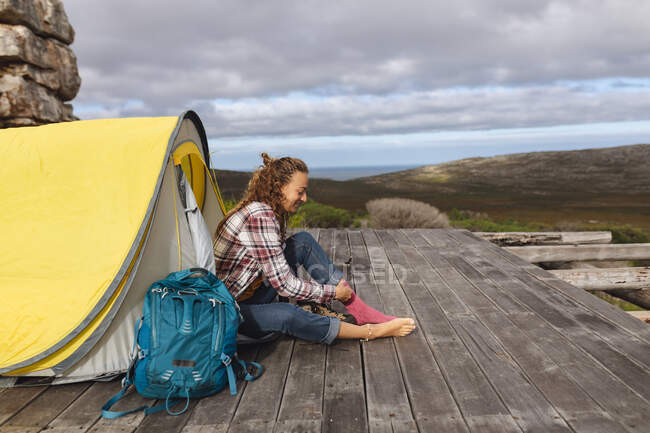 Feliz mujer caucásica acampando, sentada fuera de la tienda en la cubierta de la montaña poniéndose calcetines. vida sana, fuera de la red y cerca de la naturaleza. - foto de stock