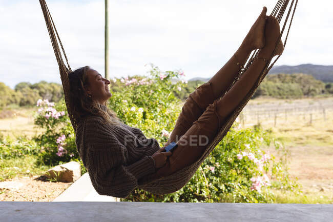 Mulher branca sorridente deitada na rede no terraço ensolarado da casa de campo, olhando para o jardim. vida saudável, perto da natureza em fora da grade casa rural. — Fotografia de Stock