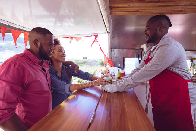 Smiling african american man in food truck chatting to male and female customers. independent business and street food service concept. — Stock Photo