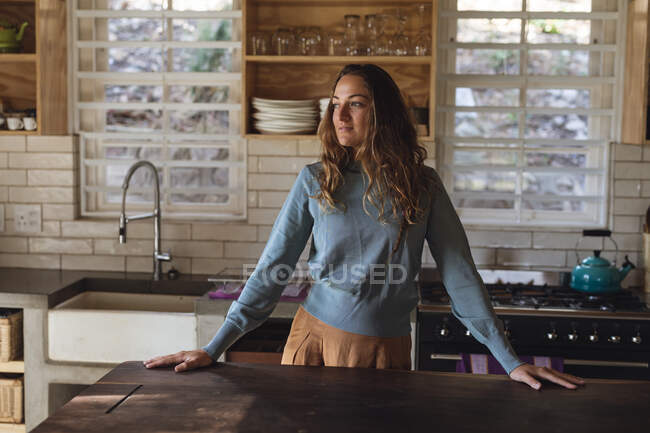 Mujer caucásica feliz de pie en la cocina de la cabaña detrás del mostrador mirando hacia otro lado. simple vivir en una casa rural fuera de la red. - foto de stock