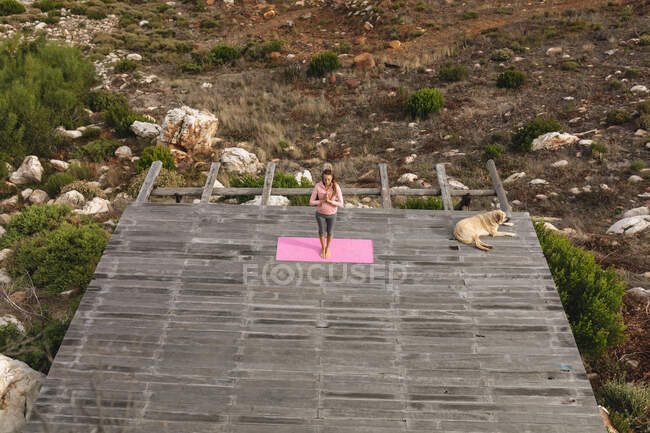 Спокойная кавказская женщина практикующая йогу стоя в медитации на палубе в сельской местности. здоровый образ жизни, вне сети и близко к природе. — стоковое фото