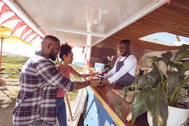 Sonriente hombre afroamericano en camión de comida charlando con clientes masculinos y femeninos. concepto de empresa independiente y servicio de comida callejera. - foto de stock