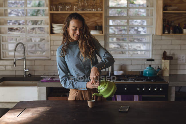 Mulher caucasiana feliz em pé na cozinha cottage derramando chá de bule e sorrindo. simples viver em uma casa rural fora da grade. — Fotografia de Stock