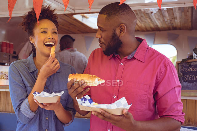 Lächelndes vielseitiges Paar, das an sonnigen Tagen Würstchen und Kartoffelkeile per Foodtruck verspeist. Unabhängiges Geschäfts- und Streetfood-Konzept. — Stockfoto