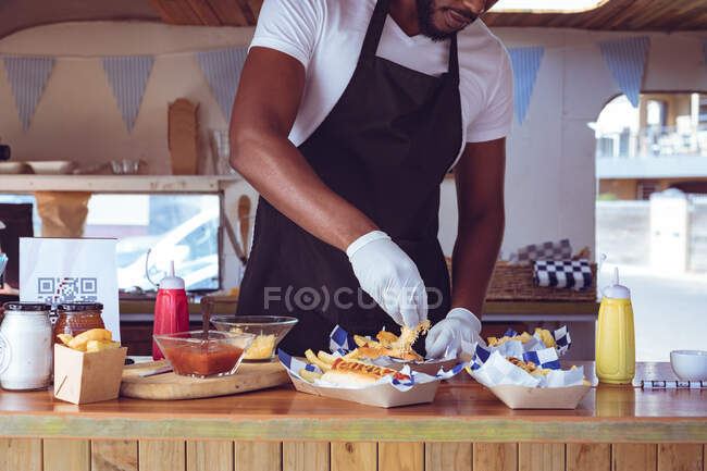 Mittelteil eines afrikanisch-amerikanischen Mannes im Foodtruck bei der Zubereitung einer Bestellung mit Hot-Dogs auf der Arbeitsplatte. Unabhängiges Geschäfts- und Streetfood-Konzept. — Stockfoto