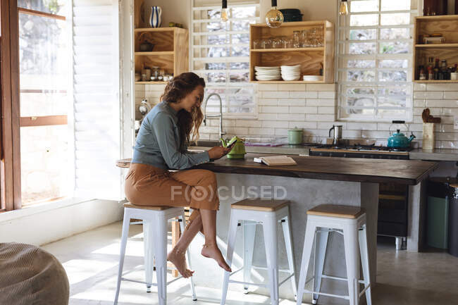 Mulher caucasiana feliz sentado no balcão na cozinha cottage usando smartphone e sorrindo. simples viver em uma casa rural fora da grade. — Fotografia de Stock
