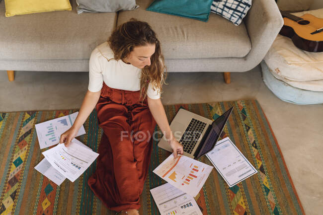 Mulher branca trabalhando em casa sentada no chão com papelada e laptop. trabalhar em casa de forma isolada durante o confinamento de quarentena. — Fotografia de Stock