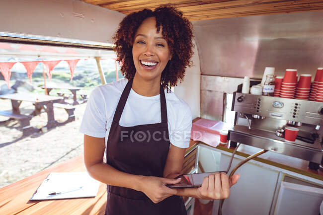Portrait of smiling mixed race woman behind counter using smartphone in food truck. independent business and street food service concept. — Stock Photo